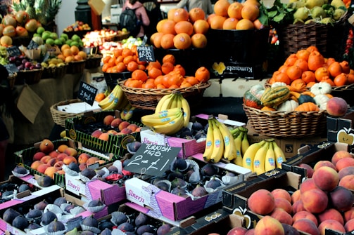 A vibrant market scene with a wide variety of fruits on display. Baskets and boxes are filled with oranges, bananas, figs, various types of squash, and peaches. Signs indicate prices and labels for the produce, creating a lively and colorful assortment.
