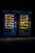Two illuminated vending machines stand side by side in a dimly lit environment. The left machine is filled with various snacks, while the right one has an array of beverage bottles. Both machines are labeled 'Grab & Go' at the top.