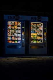 Two illuminated vending machines stand side by side in a dimly lit environment. The left machine is filled with various snacks, while the right one has an array of beverage bottles. Both machines are labeled 'Grab & Go' at the top.