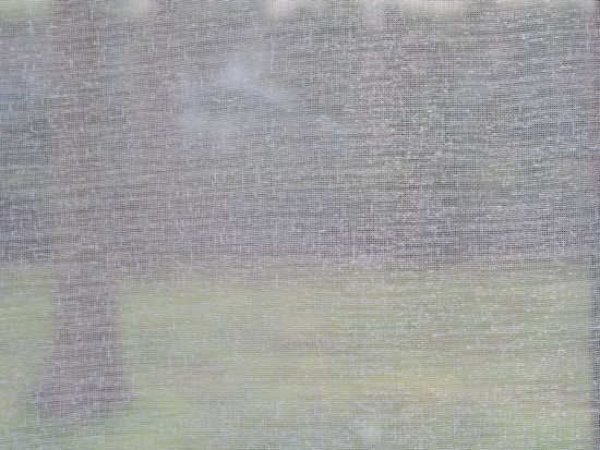 A mesh or screen material is captured up close, revealing its texture. Beyond the mesh, a blurred background suggests a natural outdoor setting, possibly with trees and grass.