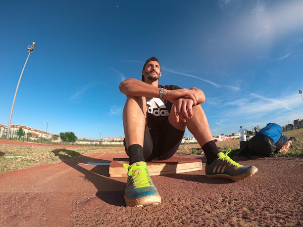 A person wearing a black athletic outfit is seated on a wooden platform in an outdoor sports field. The individual is relaxed, with arms resting on knees. The field is expansive with a clear blue sky and scattered clouds above. A brightly colored running shoe and athletic gear are visible, along with a water bottle and a blue backpack placed beside the person. Lampposts and residential buildings can be seen in the background.