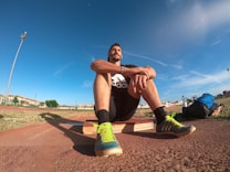 A person wearing a black athletic outfit is seated on a wooden platform in an outdoor sports field. The individual is relaxed, with arms resting on knees. The field is expansive with a clear blue sky and scattered clouds above. A brightly colored running shoe and athletic gear are visible, along with a water bottle and a blue backpack placed beside the person. Lampposts and residential buildings can be seen in the background.