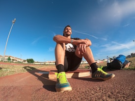 A person wearing a black athletic outfit is seated on a wooden platform in an outdoor sports field. The individual is relaxed, with arms resting on knees. The field is expansive with a clear blue sky and scattered clouds above. A brightly colored running shoe and athletic gear are visible, along with a water bottle and a blue backpack placed beside the person. Lampposts and residential buildings can be seen in the background.