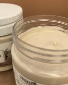 Two jars of cream or lotion with one open jar showing a smooth, creamy substance inside. The jars have white lids and label designs featuring text and botanical illustrations.