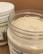 Two jars of cream or lotion with one open jar showing a smooth, creamy substance inside. The jars have white lids and label designs featuring text and botanical illustrations.