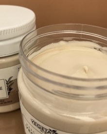Two jars of cream or lotion with one open jar showing a smooth, creamy substance inside. The jars have white lids and label designs featuring text and botanical illustrations.