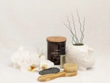 A collection of spa and relaxation items is arranged on a light surface. White orchids are placed next to a glass jar labeled Rosemary, filled with a wood wick soy candle. A small glass bottle of essential oil stands nearby. A decorative white elephant-shaped planter displays some curly green plants. In the foreground, a natural bristle body brush and a smooth black stone complete the arrangement.