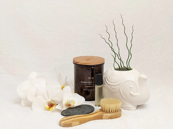 A collection of spa and relaxation items is arranged on a light surface. White orchids are placed next to a glass jar labeled Rosemary, filled with a wood wick soy candle. A small glass bottle of essential oil stands nearby. A decorative white elephant-shaped planter displays some curly green plants. In the foreground, a natural bristle body brush and a smooth black stone complete the arrangement.
