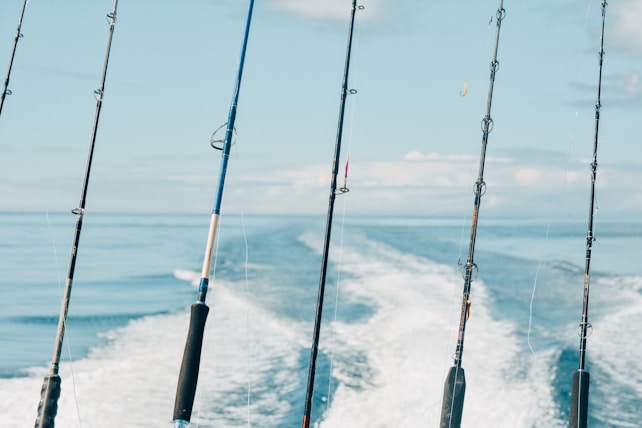 Several fishing rods are secured on a boat, positioned as the vessel moves swiftly across a calm body of water, creating foamy wake trails in the background against a serene blue sky.