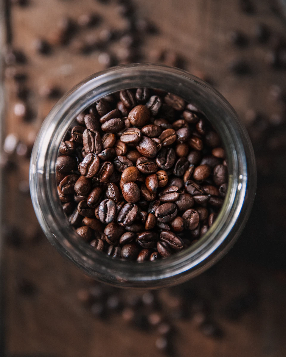 The image shows a glass jar filled with coffee beans, placed on a wooden surface. The jar is transparent and allows the viewer to see the beans inside. The beans are dark brown in color and appear to be freshly roasted. They are scattered throughout the jar, some overlapping each other.