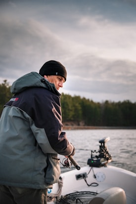 A person dressed warmly stands on a boat holding a fishing rod against a backdrop of a lake and trees. The cloudy sky suggests an overcast day, and the equipment on the boat is visible, hinting at a fishing expedition.