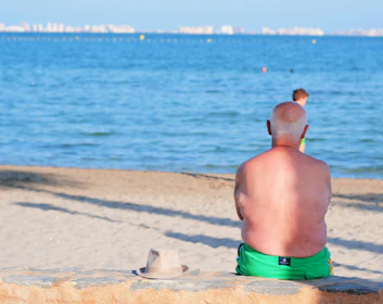 A shirtless, elderly man with short hair is sitting with his back to the camera on a stone ledge by the beach. He is wearing bright green swim trunks. A straw hat is placed beside him on the ledge. The man is gazing towards the ocean where a few people and distant buildings are visible across the water.