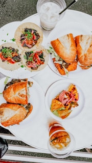 An assortment of food items is presented on white disposable plates placed on a round table. The spread includes three small tacos topped with savory ingredients, two pairs of sandwiches using crusty bread filled with meats or vegetables, and a small plate with a slice of quiche or frittata garnished with pickled onions. Accompanying the meal are two beverages, one a milky drink with ice and the other a dark iced coffee.