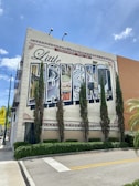 A large mural on the side of a building depicts a collage of culturally significant images, with the word 'Havana' prominently displayed. Tall, thin evergreen trees line the front of the building, and the sky is bright and clear with a few clouds. The scene includes palm trees and a street sign indicating a pedestrian crossing.