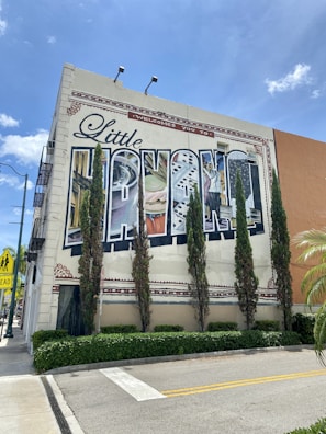 A large mural on the side of a building depicts a collage of culturally significant images, with the word 'Havana' prominently displayed. Tall, thin evergreen trees line the front of the building, and the sky is bright and clear with a few clouds. The scene includes palm trees and a street sign indicating a pedestrian crossing.