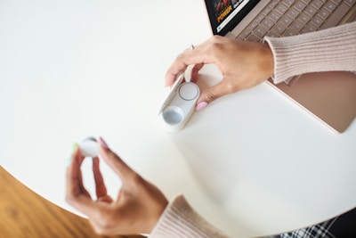 Hands of a person with painted nails are holding a small white gadget on a white surface near a laptop. The person seems to be interacting with the gadget, possibly inserting or removing a component.