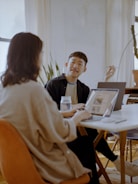 Two people are sitting at a table in a well-lit room with laptops open in front of them. One person is gesturing with a pen, possibly explaining something, while the other listens intently. There is a jar on the table and plants in the background.