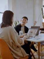 Two people are sitting at a table in a well-lit room with laptops open in front of them. One person is gesturing with a pen, possibly explaining something, while the other listens intently. There is a jar on the table and plants in the background.