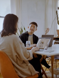 Two people are sitting at a table in a well-lit room with laptops open in front of them. One person is gesturing with a pen, possibly explaining something, while the other listens intently. There is a jar on the table and plants in the background.