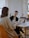 Two people are sitting at a table in a well-lit room with laptops open in front of them. One person is gesturing with a pen, possibly explaining something, while the other listens intently. There is a jar on the table and plants in the background.