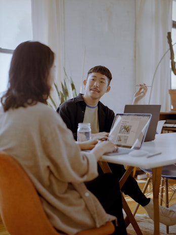 Two people are sitting at a table in a well-lit room with laptops open in front of them. One person is gesturing with a pen, possibly explaining something, while the other listens intently. There is a jar on the table and plants in the background.