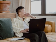 man in white dress shirt and black pants sitting on couch using Surface laptop