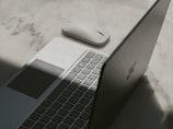 A laptop with a partially open lid beside a wireless mouse placed on a textured surface. The lighting casts a shadow across the laptop, emphasizing its sleek design and minimalist features.