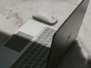 A laptop with a partially open lid beside a wireless mouse placed on a textured surface. The lighting casts a shadow across the laptop, emphasizing its sleek design and minimalist features.