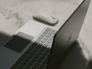 A laptop with a partially open lid beside a wireless mouse placed on a textured surface. The lighting casts a shadow across the laptop, emphasizing its sleek design and minimalist features.