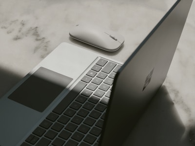 A laptop with a partially open lid beside a wireless mouse placed on a textured surface. The lighting casts a shadow across the laptop, emphasizing its sleek design and minimalist features.