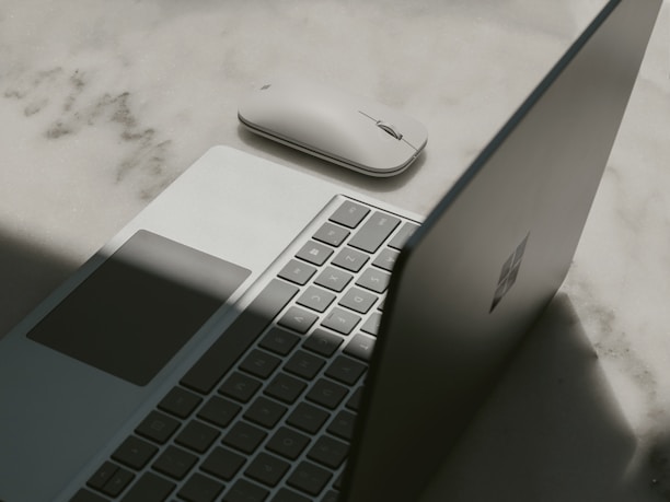 A laptop with a partially open lid beside a wireless mouse placed on a textured surface. The lighting casts a shadow across the laptop, emphasizing its sleek design and minimalist features.
