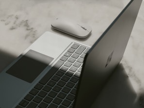 A laptop with a partially open lid beside a wireless mouse placed on a textured surface. The lighting casts a shadow across the laptop, emphasizing its sleek design and minimalist features.