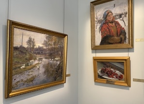 The image features a corner of an art gallery displaying three paintings. The left painting depicts a serene landscape with a river, trees, and a distant bridge. The top right painting shows a woman wearing a traditional attire with a red scarf, blue headscarf, and several necklaces, standing against a snowy background. The bottom right painting is a still life of a plate with red berries.