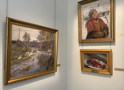 The image features a corner of an art gallery displaying three paintings. The left painting depicts a serene landscape with a river, trees, and a distant bridge. The top right painting shows a woman wearing a traditional attire with a red scarf, blue headscarf, and several necklaces, standing against a snowy background. The bottom right painting is a still life of a plate with red berries.