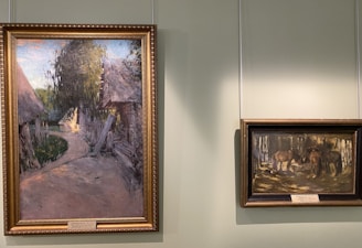 Two framed paintings are displayed on a light green wall. The larger painting on the left depicts a rural scene with a path leading through a village surrounded by trees and traditional thatched-roof huts. The smaller painting on the right shows a group of horses standing together in a wooded area.