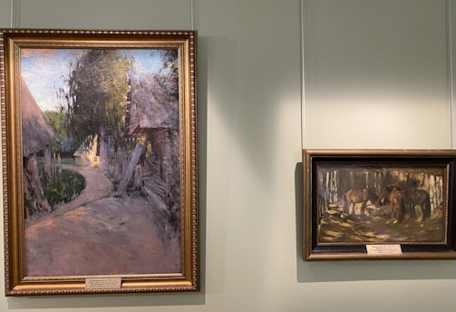 Two framed paintings are displayed on a light green wall. The larger painting on the left depicts a rural scene with a path leading through a village surrounded by trees and traditional thatched-roof huts. The smaller painting on the right shows a group of horses standing together in a wooded area.