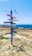 A colorful signpost stands on rocky terrain near a coastline, pointing to various global destinations, with distances written on each directional plank. The ocean is visible in the background, meeting a clear blue sky.