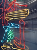 Neon lights in various colors, including red, blue, and yellow, are arranged in a complex design against a corrugated metal background. The lights form abstract shapes and patterns, creating a vibrant and dynamic visual effect.