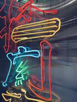 Neon lights in various colors, including red, blue, and yellow, are arranged in a complex design against a corrugated metal background. The lights form abstract shapes and patterns, creating a vibrant and dynamic visual effect.
