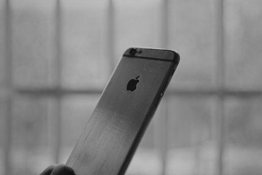A close-up, monochrome image of the back of a smartphone with the logo visible. The phone is held by a hand, and the background shows blurred vertical and horizontal lines, suggesting a window or bars.