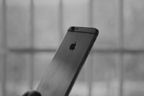 A close-up, monochrome image of the back of a smartphone with the logo visible. The phone is held by a hand, and the background shows blurred vertical and horizontal lines, suggesting a window or bars.