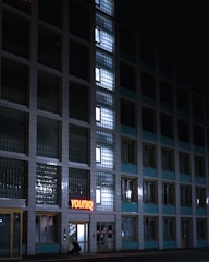 A multi-story building with large, grid-like glass windows lit from within, creating a pattern of light and shadow. A neon sign reading 'YOUNIQ' is displayed near the entrance. A person is crouched near the doorway, possibly engaged in some activity. The overall scene is dimly lit, suggesting nighttime.