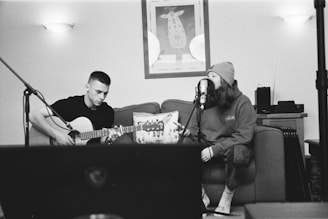 A black and white photograph of two people in a cozy indoor setting. One person is playing an acoustic guitar, while the other appears to be singing into a microphone, seated on a couch. A wall with framed art and soft lighting create a relaxed atmosphere.