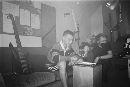 Two individuals are seated in a dimly lit room. One person is interacting with a digital music controller placed on a small table in front of them, while the other watches attentively. A guitar leans against the sofa, and a bulletin board with papers is visible on the wall. Various equipment, including a camera, is in the surroundings, suggesting a creative setting.
