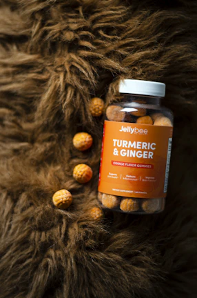 A jar of turmeric and ginger orange flavor gummies is placed on a textured brown surface, with a few gummies scattered around. The jar is clear with an orange label, featuring the brand name 'Jellybee'. The surface resembles fur, adding a cozy and natural element to the composition.