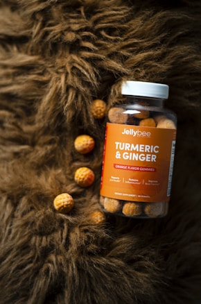 A jar of turmeric and ginger orange flavor gummies is placed on a textured brown surface, with a few gummies scattered around. The jar is clear with an orange label, featuring the brand name 'Jellybee'. The surface resembles fur, adding a cozy and natural element to the composition.