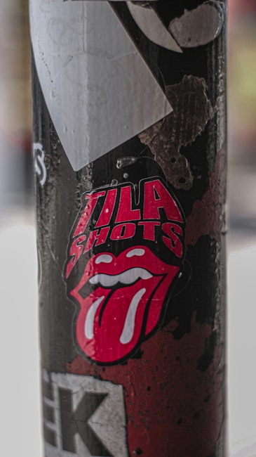 A street pole is covered with various stickers and peeling paper. Prominent among the stickers is a graphic image of a red tongue sticking out from the lips, accompanied by the words 'illa shots'. The pole shows signs of weathering and wear.