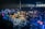 A large crowd of people gathered at a concert or festival, with vibrant lighting and lively atmosphere. Attendees are enthusiastically raising their hands, taking photos, and enjoying the performance. A metal barrier separates the audience from the stage area.