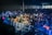 A large crowd of people gathered at a concert or festival, with vibrant lighting and lively atmosphere. Attendees are enthusiastically raising their hands, taking photos, and enjoying the performance. A metal barrier separates the audience from the stage area.
