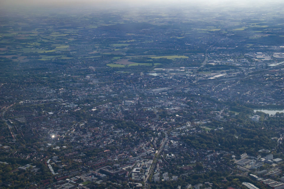 The image presents an aerial view of a city, captured from above and bathed in soft light. The cityscape is densely populated with buildings of varying heights, creating a sense of depth and scale. A network of roads crisscrosses the landscape, connecting different parts of the city. The sky above is a uniform blue color, providing a stark contrast to the urban environment below. The image does not contain any discernible text or countable objects, but it does provide a comprehensive view of the city's layout and infrastructure.